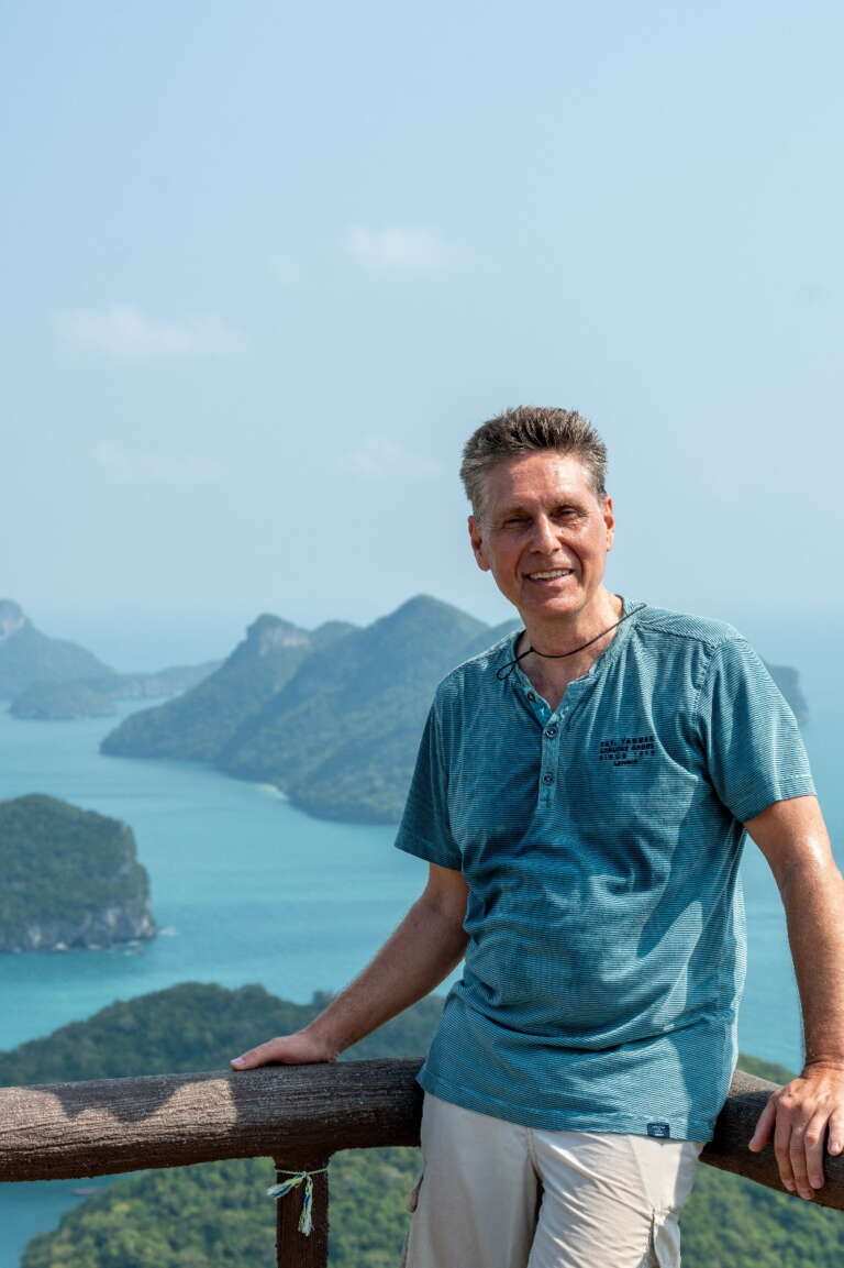 Lotus Vita founder Michael Wolf stands smiling on a viewpoint, with a scenic background of tropical islands and turquoise water.