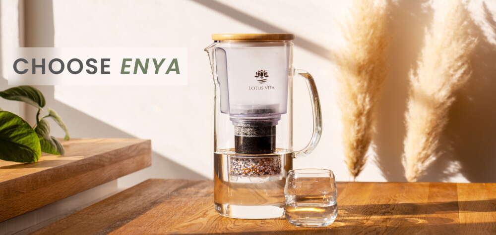 The Lotus Vita ENYA water filter pitcher, made of glass with a bamboo lid, sits on a sunlit wooden table next to a glass of fresh water.