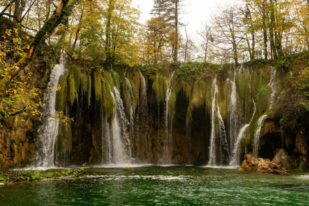 A wide, multi-stream waterfall flows over a moss-covered cliff into a clear, green pool, embodying the purity of nature.